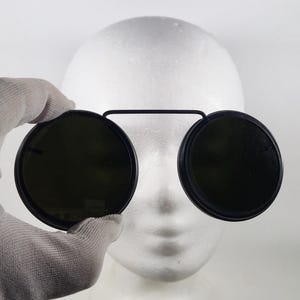 Può includere: Occhiali da sole rotondi con lenti verde scuro e montatura nera. Le lenti sono collegate da un ponte nero. Gli occhiali da sole sono tenuti davanti a una testa di manichino bianca.