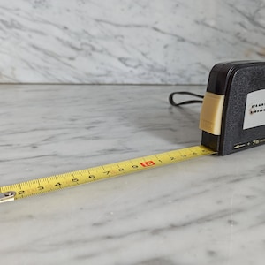 Könnte beinhalten: Ein gelbes Maßband mit schwarzen Markierungen erstreckt sich von einem schwarzen Maßband mit einem weißen Knopf und dem Text "PLASSER TRIMMER" auf der Vorderseite. Das Maßband liegt auf einer weißen Marmorfläche.