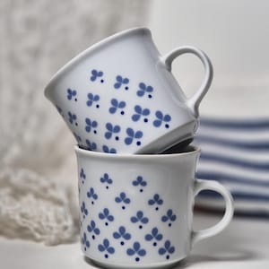 Peut inclure: Deux tasses en céramique blanche empilées verticalement, chacune présentant un motif répétitif de petits trèfles bleus à trois feuilles et de minuscules points bleus. Les tasses ont une finition lisse, une anse arrondie et un bord légèrement évasé.