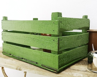 May include: A green wooden crate with a rustic finish. The crate has three horizontal slats and is sitting on a wooden surface.