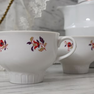 Può includere: Due tazze da tè bianche con motivi floreali in rosso, oro e viola. Le tazze hanno una base larga e un bordo svasato.