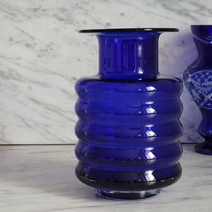 May include: A cobalt blue glass vase with a ribbed design. The vase has a round base and a narrow neck.