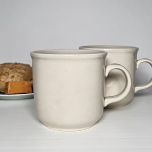 Peut inclure: Deux tasses en céramique blanc cassé avec une finition lisse et mouchetée et une anse arrondie. Les tasses présentent une forme cylindrique simple avec un bord légèrement évasé, offrant une esthétique propre et neutre pour servir des boissons.