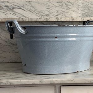 Könnte beinhalten: Eine hellblaue Emaille-Waschwanne mit zwei Metallgriffen. Die Wanne steht auf einer weißen Marmorfläche.