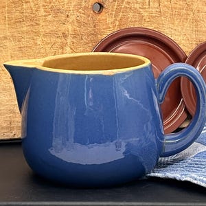 Könnte beinhalten: Ein glänzender blauer Keramikkrug mit breitem Ausguss und geschwungenem Henkel. Das Innere des Kruges ist hellbraun. Die Außenfläche hat eine glatte, reflektierende Oberfläche mit einer kleinen Absplitterung am oberen Rand des Gefäßes.