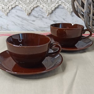 May include: Two brown ceramic teacups and saucers. The cups have a rounded shape and a smooth, glossy finish. The saucers are slightly larger than the cups and have a matching brown colour.