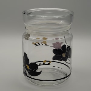 Op de afbeelding: Een glazen pot met een zwart en gouden bloemenmotief. De pot heeft een deksel en is perfect voor het bewaren van kleine voorwerpen.