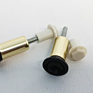 Könnte beinhalten: Zwei goldene Möbelbeinabdeckungen aus Metall mit schwarzen Kunststoff Füßen und weißen Kunststoff Schraubkappen. Die Abdeckungen sind so konzipiert, dass sie an Möbelbeinen befestigt werden können.