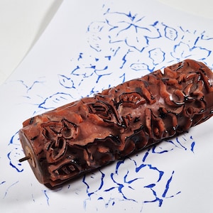 May include: A cylindrical brown rubber paint roller with a raised floral pattern. The roller is positioned on a white sheet of paper that displays a blue ink print of the same floral design, showing the texture and pattern created by the tool.