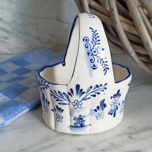 Könnte beinhalten: Ein weißer Keramikkorb mit blau-weißem Delftware-Design. Der Korb hat einen Griff und ist mit Blumen und einer Windmühle verziert.