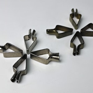 Op de afbeelding: Een set van acht metalen fotohaakjes in zilver en goud. De haakjes zijn driehoekig met een klein haakje aan de bovenkant.