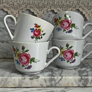 Op de afbeelding: Vier witte keramische theekopjes gestapeld op een marmeren oppervlak. Elke kop heeft een grote roze roos met groene bladeren en kleinere kleurrijke bloemen. De kopjes hebben gebogen handvatten.
