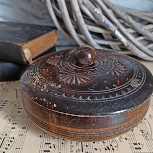 May include: A dark brown wooden box with a carved design and a lid. The box has a small knob on the top of the lid.