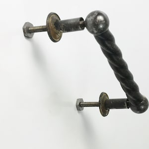 May include: A black metal door handle with a twisted design and round ends. The handle is attached to a wall with two screws and metal plates.