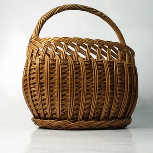 May include: A brown wicker basket with a curved handle. The basket has a rounded body and a braided base. The handle is made of woven wicker. The basket is designed for carrying items. The basket is made of woven wicker.