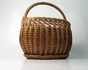 May include: A woven wicker basket with a rounded body and a curved handle. The basket is made of light brown wicker and has a braided base. The handle is also made of woven wicker. The basket is designed for carrying items.