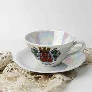 May include: A white teacup and saucer with a pearl-like iridescent glaze. The teacup has a handle and features a colourful coat of arms with a castle and the text "Munchen Deutschland".