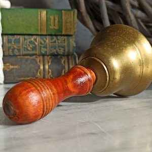 Può includere: Una campana di ottone con un manico di legno. La campana è dorata e ha una forma arrotondata. Il manico è marrone e ha una superficie testurizzata.