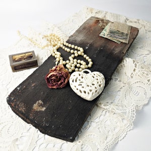 May include: A white ceramic heart pendant with a delicate floral design, a string of white pearls, and a dried rose on a dark brown wooden surface.