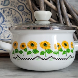Op de afbeelding: Een witte emaille pan met een zonnebloemdessin en een zwart-wit geblokte rand. De pan heeft een zilveren deksel met een witte knop.
