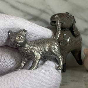 Op de afbeelding: Een zilverkleurig kattenbeeldje met gedetailleerde vachttextuur, naast een hondenbeeldje. De kat heeft een gefocuste blik en een opgetrokken staart. De hond heeft een afgerond lichaam en een kleine staart. Een klein roze en wit object staat op de achtergrond.