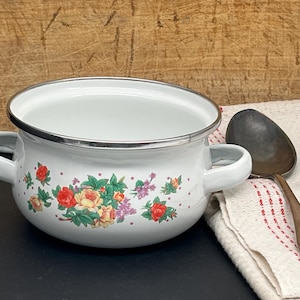 Op de afbeelding: Een witte emaille pan met twee zijhandvatten en een zilveren rand, voorzien van een bloemmotief van rode en gele rozen met groene bladeren en kleine paarse bloemetjes, vergezeld door een donkere metalen soeplepel op een witte doek.