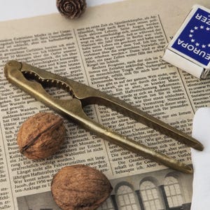 May include: A vintage brass nutcracker with a walnut and two halves of a walnut on a newspaper. A matchbox with "EUROPA" printed on it is also visible.
