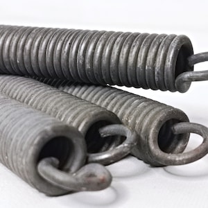May include: Close-up of several industrial-grade metal springs. The springs are a dark gray color, with a coiled design and curved end loops. The image is well-lit, highlighting the texture and form of the springs.
