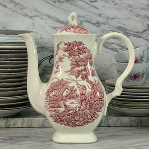 May include: A white ceramic coffee pot with a pink floral design. The design features a hunting scene with a man and a dog in a woodland setting. The pot has a lid and a handle.