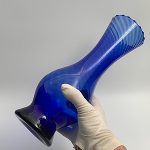 Peut inclure: Un vase en verre bleu, élégant et élancé, avec une base évasée et un bord ondulé. Le vase présente une forme incurvée et allongée, avec un dégradé de bleus, du bleu indigo profond aux nuances plus claires. Le vase est tenu par une main gantée.