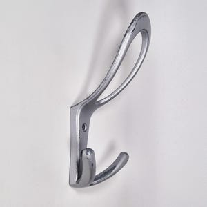 May include: A silver-colored metal wall hook with a curved design. The hook has a rectangular base with a single hook and a curved top loop. The metal has a distressed finish, showing signs of wear.