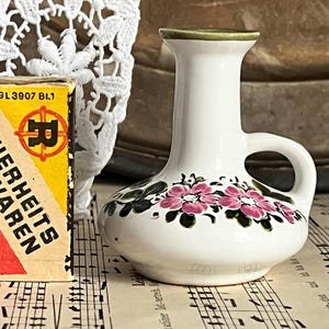 Könnte beinhalten: Kleine, weiße Keramikvase mit Henkel, verziert mit rosa Blumendesigns und grünen Blättern. Die Vase hat einen grünen Rand und einen abgerundeten Boden. Eine Vintage-Streichholzschachtel mit deutschem Text befindet sich daneben.