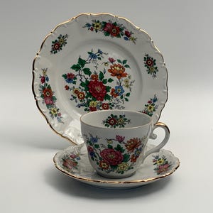 Può includere: Un servizio da tè in porcellana vintage con una tazza, un piattino e un piatto da portata. Il set è bianco con finiture dorate e un motivo floreale rosso, arancione, rosa e blu. Il piatto ha un bordo smerlato.