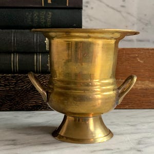 Könnte beinhalten: Eine kleine, goldfarbene Metallvase mit zwei Henkeln. Die Vase hat einen runden Boden und einen zylindrischen Körper. Sie steht auf einer weißen Marmorfläche.