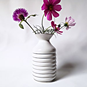 Könnte beinhalten: Eine weiße Keramikvase mit geripptem Design hält einen Strauß rosa und lila Blumen. Die Vase hat einen abgerundeten Boden und einen ausgestellten oberen Rand.