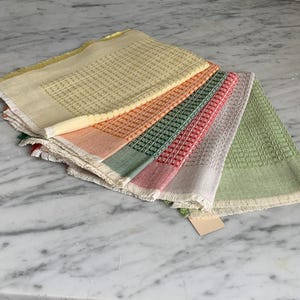 May include: A stack of colorful, textured kitchen towels in shades of yellow, orange, green, pink, and gray. Each towel has a woven pattern and a fringed edge. The towels are neatly arranged, showcasing their varied colors.