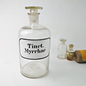 May include: A collection of vintage apothecary bottles. The largest bottle has a label that reads "Tinct. Myrrhae." Other bottles and a spool of twine with a yellow label are also present. The bottles are clear glass.