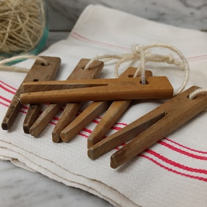 May include: Several vintage wooden clothes pegs with string attached, displayed on a white cloth with red stripes. The pegs are made of light brown wood and have a traditional design. A ball of twine is visible in the background.