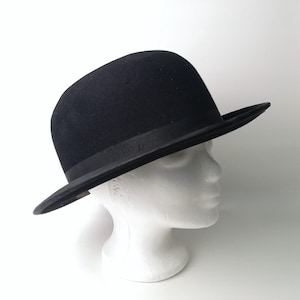 May include: A black bowler hat with a wide brim. The hat is sitting on a white mannequin head.