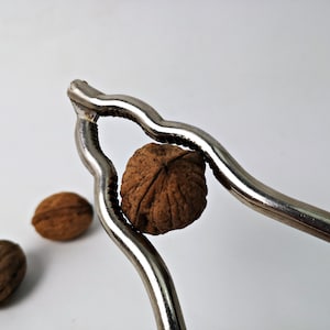 Könnte beinhalten: Ein silberner Nussknacker mit einer braunen Walnuss in den Backen. Zwei weitere Walnüsse liegen auf der weißen Oberfläche.