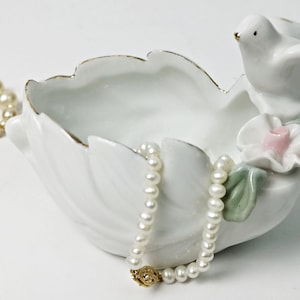 Può includere: Piatto portaoggetti in porcellana bianca a forma di uccello con un fiore e foglie. Il piatto ha un bordo dorato ed è decorato con un fiore rosa. Due collane di perle sono nel piatto.