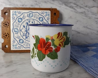 May include: White enamel mug with a blue rim and a floral design featuring red, yellow, and green flowers. The mug is sitting on a marble surface.