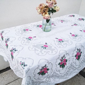 May include: White tablecloth with a gray floral pattern and pink roses. The tablecloth is laid out on a table with a vase of dried flowers in the center.