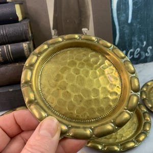 May include: Set of four brass coasters with a hammered finish and a decorative rim. The coasters are round and have a slightly raised edge.