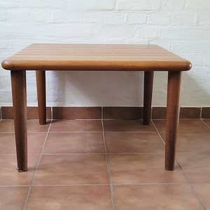 Könnte beinhalten: Ein quadratischer Holz-Couchtisch mit abgerundeten Kanten und vier Beinen. Der Tisch ist aus hellbraunem Holz gefertigt und hat eine natürliche Oberfläche.