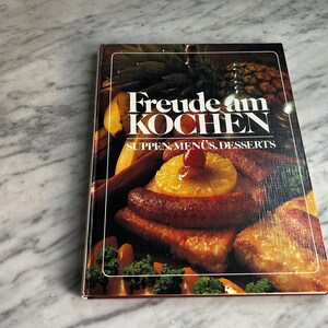 May include: A hardback cookbook titled Freude am KOCHEN SUPPEN, MENÜS, DESSERTS. The cover features a photograph of a cooked dish with pineapple slices, sausages, and a cherry, alongside a pineapple, all set against a dark background.