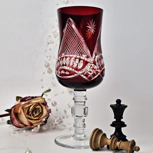 May include: A red crystal wine glass with white etched patterns. A dried rose, a pearl necklace, and chess pieces are arranged near the glass. The chess pieces include a black queen and a fallen beige king.
