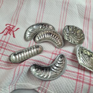 以下が含まれることがあります： 三日月形と長方形の銀色の金属製の焼き型が6つ。焼き型は白と赤の市松模様の布の上に置かれています。