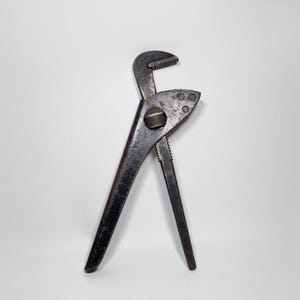May include: A well-worn, dark metal pipe wrench. The adjustable jaws are open, revealing the teeth designed to grip pipes. The tool has a pivot bolt and a textured handle for a secure grip. The wrench is set against a white background.