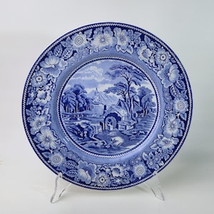 Op de afbeelding: Een blauw-wit decoratief bord met een bloemenrand en een centrale landschapsscène. Het bord wordt op een transparante standaard gepresenteerd. Het landschap toont bomen, een gebouw en een brug.